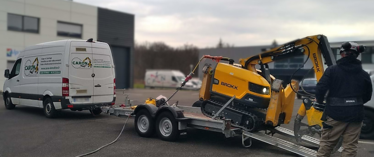 Un fourgon Caron Démolition est stationné. Il y une remorque porte engin attelée au fourgon avec un robot de démolition dessus. L'opérateur Caron Démolition est en train de téléguidé le robot pour le descendre de la remorque porte engin. Le robot est radiocommandé à distance. Le robot de démolition est un BROKK 170.