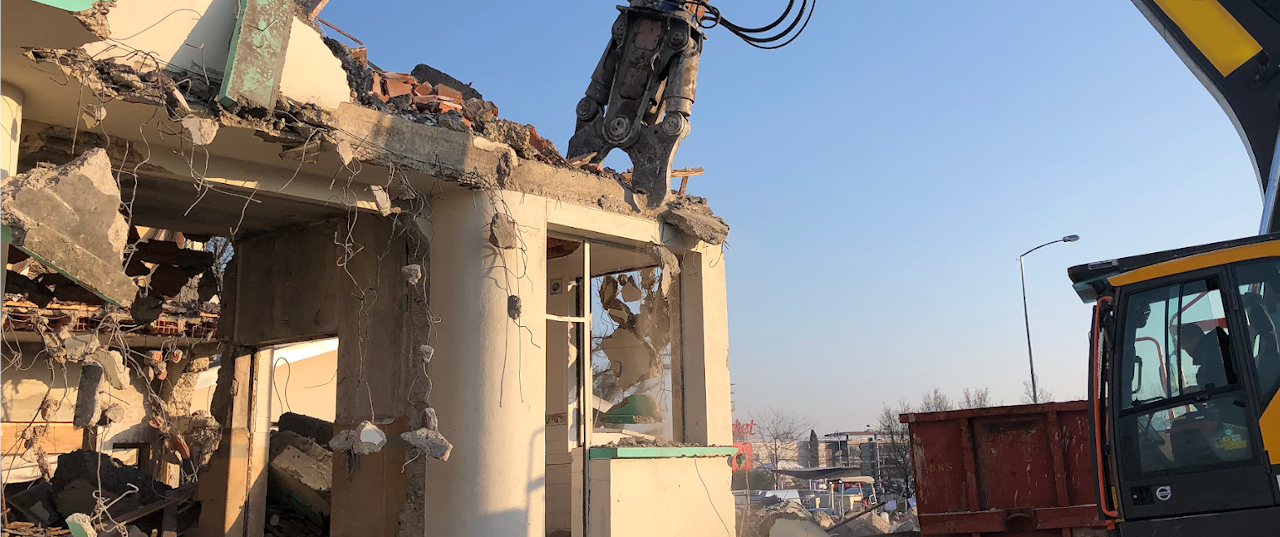 Sur la photo, nous voyons une pelleteuse en train de grignoter un bâtiment. A gauche de la photo, nous voyons le bâtiment déjà un peu démoli. A droite, nous voyons le corps de la pelleteuse avec l'opérateur dedans. En arrière plan, le ciel est bleu. La pelle est munie d'un cisaille à béton et elle est en train de coquer la poutre en béton située au dessus de la fenêtre.