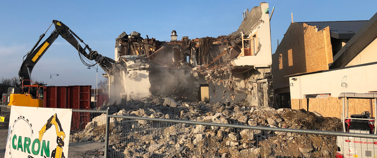 Sur la photo, au premier plan il y a une barrière de chantier avec un panneau de chantier CARON Démolition. Au centre de la photo, il y a une démolition de bâtiment en cours. La démolition du bâtiment est réalisée par une pelle 25 tonnes. Cette pelle est à gauche de la photo. Elle est en train de grignoter le mur à gauche de la bâtisse. A droite de la photo, il y a un bâtiment neuf protégé par des plaques de bois OSB..